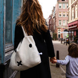 A woman with long hair and a black coat walks down the street, holding hands with a child. She proudly carries her sustainable fashion staple, the Miffy Shopping Bag from Miffy, which is crafted from recycled soft terry and showcases an adorable face design.