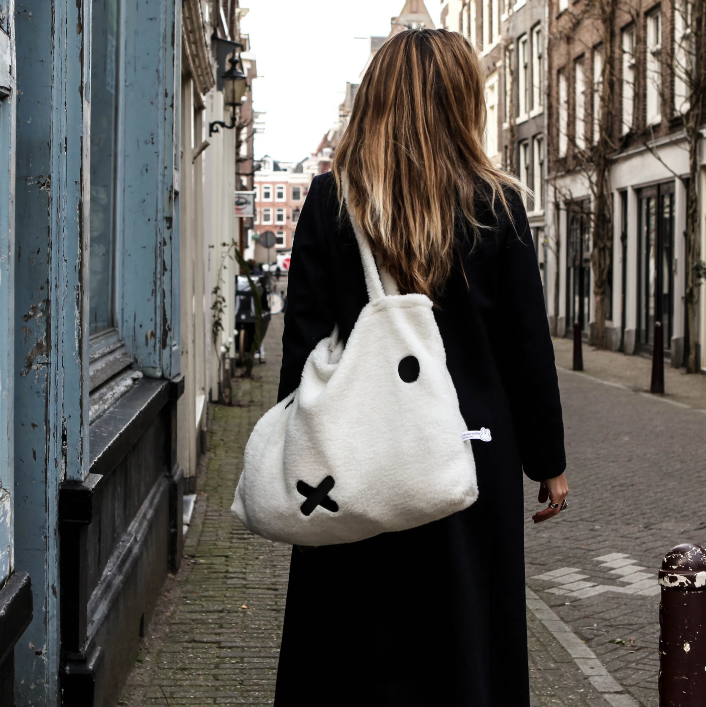 A person with long hair strolls down the street, clad in a black coat and carrying a large white Miffy Shopping Bag, crafted from recycled soft terry material by Miffy, showcasing a minimalist face design.