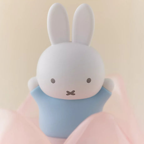 Miffy figure in light blue outfit with white ears surrounded by soft pink fabric on beige background