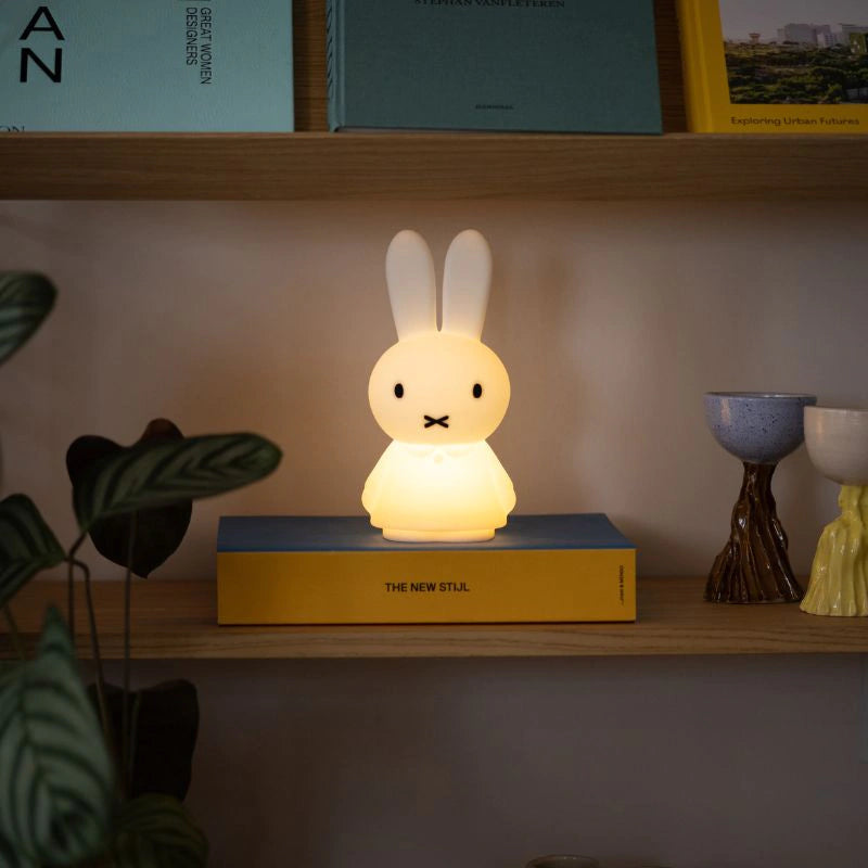 The 22cm Miffy Shines - Night Light by Miffy, with a touch-sensitive dimming feature, whimsically rests on a yellow book on the wooden shelf. It's surrounded by books, a lush plant, and charming decor, creating an inviting and cozy ambiance.