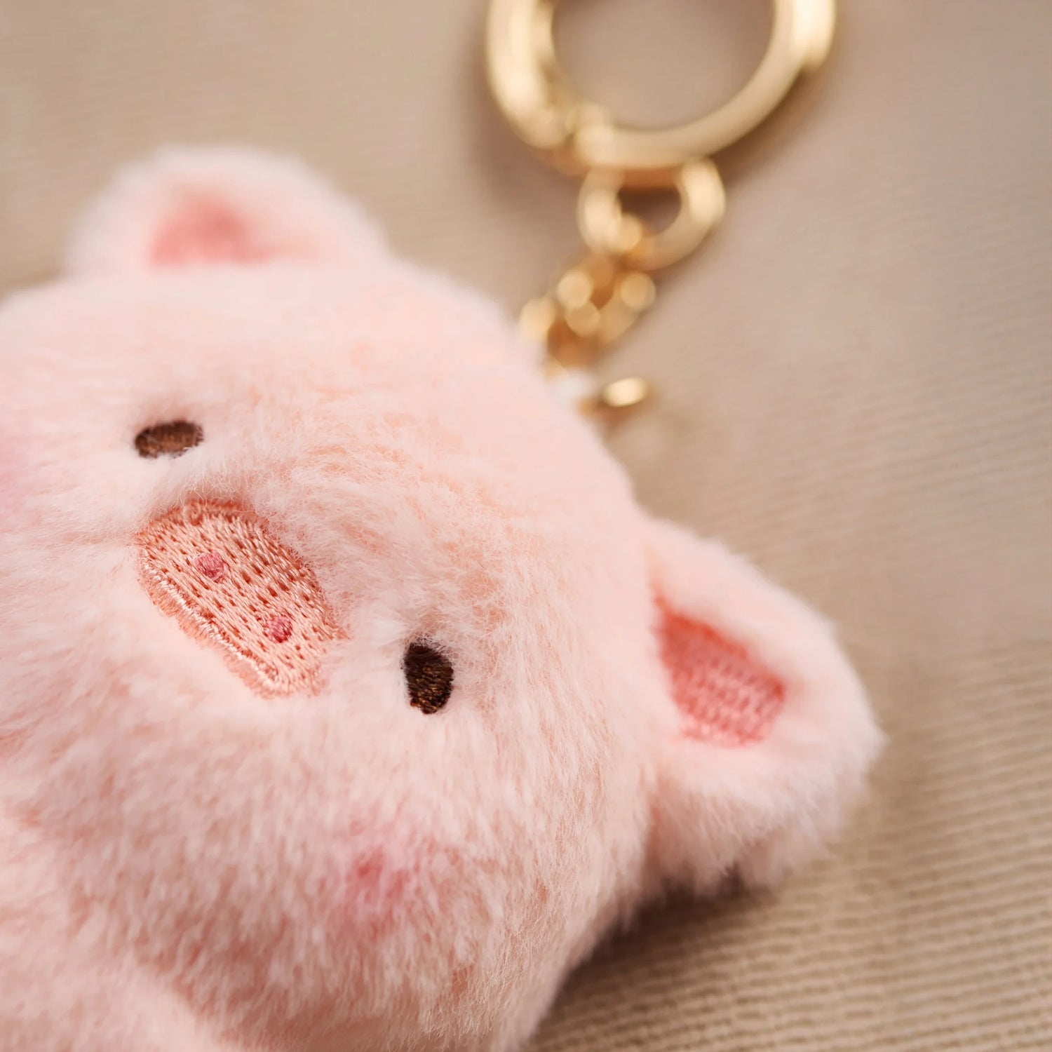 Close-up of the LuLu the Piggy - Mini LuLu Keychain by LuLu The Piggy, a soft pink pig-shaped keychain with a gold keyring, resting on a beige surface.