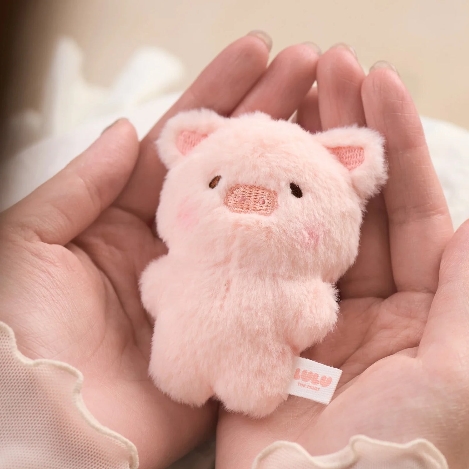 A pair of hands gently holds the LuLu The Piggy - Mini LuLu Keychain, a small plush pink pig toy with a "LULU" tag, capturing the adorable charm of LuLu the Piggy by LuLu The Piggy.