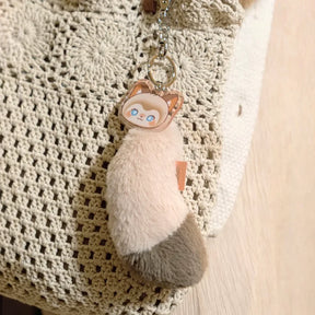 Fluffy two-toned cat tail keychain with a character charm, attached to a beige crocheted bag handle