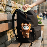 Mofusand Red Panda Mini Bag - Plush Keychain hanging from black backpack outdoors with stone wall background and hand placing wallet
