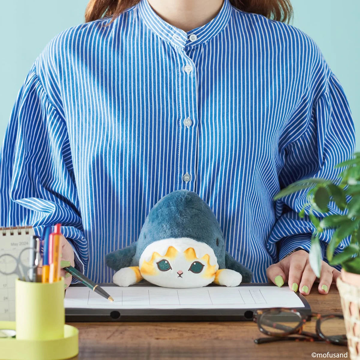 A person in a blue striped shirt sits at a desk with the Mofusand Posture Plush Buddy - Shark by Mofusand placed on a desk calendar.