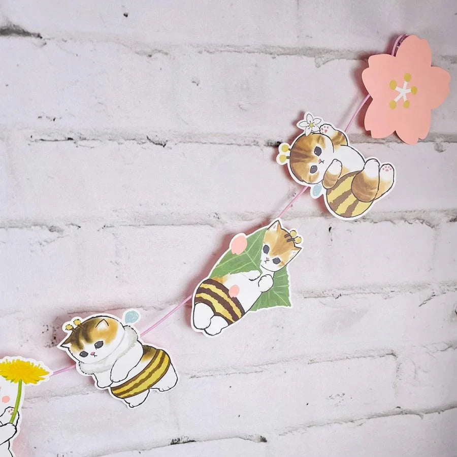 The Mofusand Spring Garland Decorations feature cartoon cat cutouts dressed as bees, with flowers and pink cherry blossoms against a white brick wall. Inspired by the whimsical Mofusand style, these playful pieces bring joy to any room.