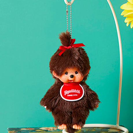 Fluffy Monchhichi Girl bag charm with brown fur, red bow on top, and red bib hanging from silver ball chain keychain