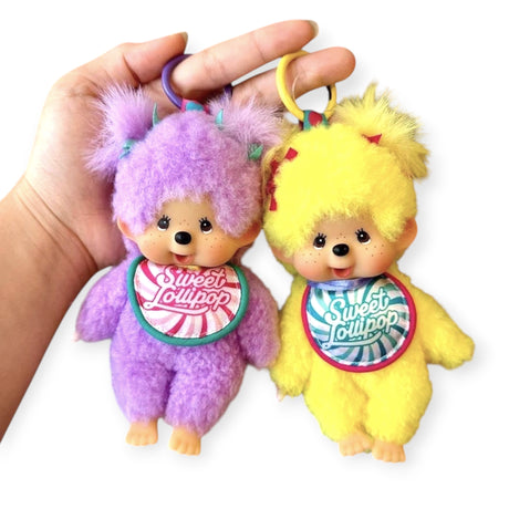 Hand holding two Monchhichi Sweet Lollipop bag charms in purple and yellow with fluffy fur and bibs hanging from colorful keyrings