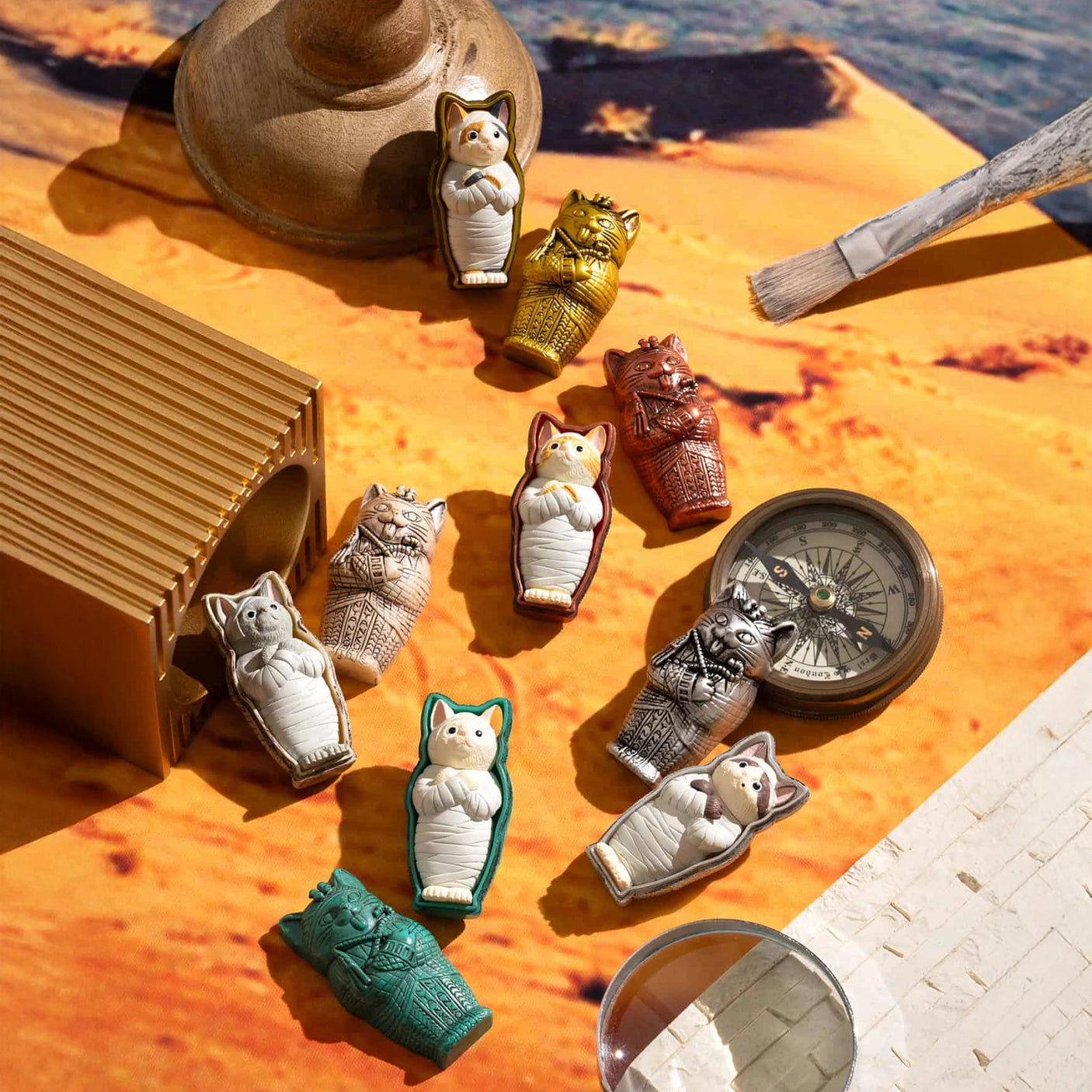 Kitan Club's Mummy Cat Blind Box figurines are showcased on a sunlit table alongside a compass, brush, and wooden object.