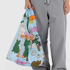 Person holding a blue bag with colorful cat illustrations, standing in gray pants.