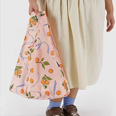 Person holding a peach-colored bag with fruit and ribbon patterns, wearing a cream skirt, blue socks, and brown shoes.