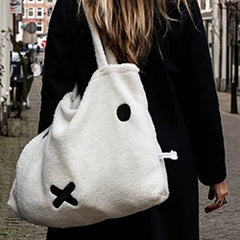 A person with long hair is walking down a street, carrying a large white bag featuring a simple black face design.