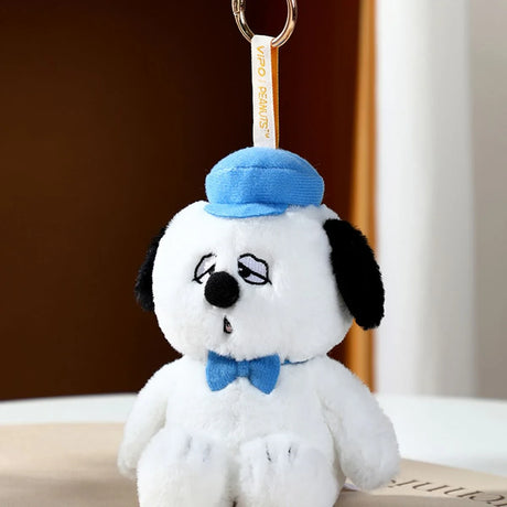 Plush toy keychain of white dog wearing blue hat and bow tie with tired eyes hanging from gold ring on neutral background
