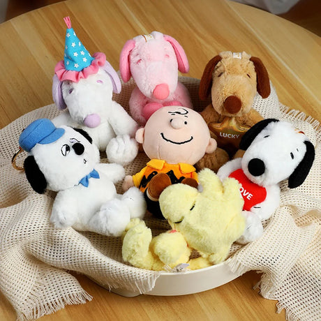 Seven Peanuts character plush toys including Charlie Brown, Snoopy, Woodstock, and others arranged on beige cloth in white bowl