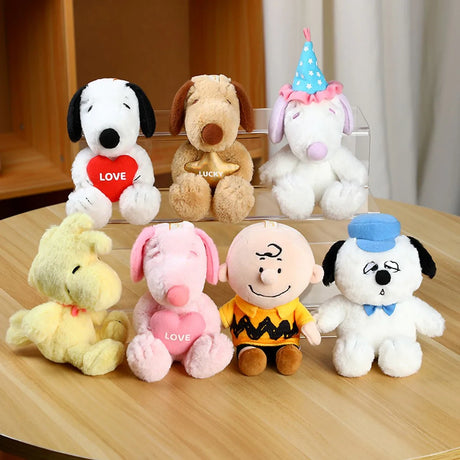 Seven Peanuts character plush toys including Snoopy, Woodstock, Charlie Brown, and others on wooden table and clear stand