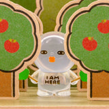 Clear-sleeve blind-box figurine standing between green hedges with orange emblem.