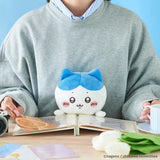 A person in a gray sweatshirt sits at a table with an open book, holding the Chiikawa Posture Plush Buddy - Hachiware by Chiikawa. A cup and camera are also on the table.