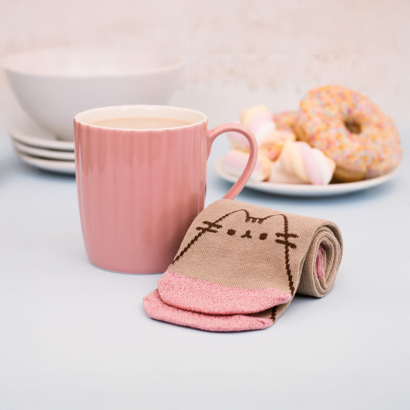 On the table, a Pusheen Sock in a Mug Gift Set showcases its ceramic pink mug and cozy socks with a cat design. In the background, there's a plate with donuts and marshmallows next to neatly stacked white bowls, creating an adorable scene.