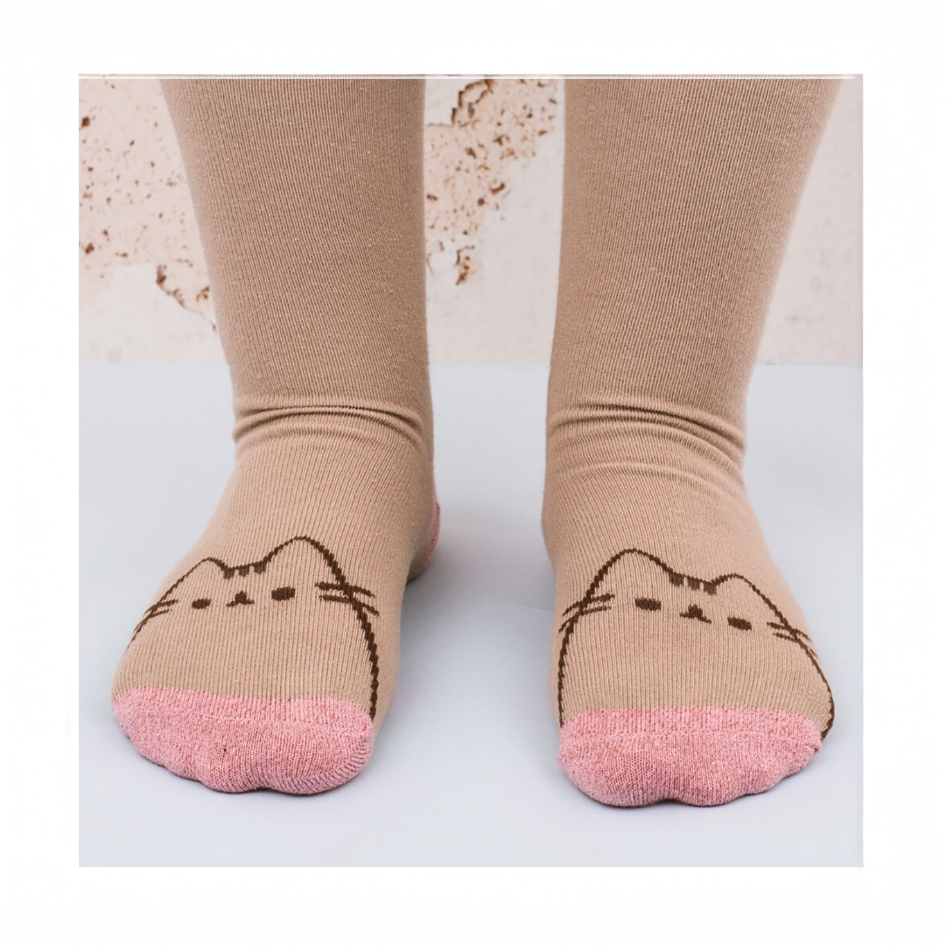 The Pusheen Sock in a Mug Gift Set features beige cozy socks with pink toes, adorned with a charming cat face design. They're worn by someone standing on a light surface, providing both warmth and whimsy to any day.