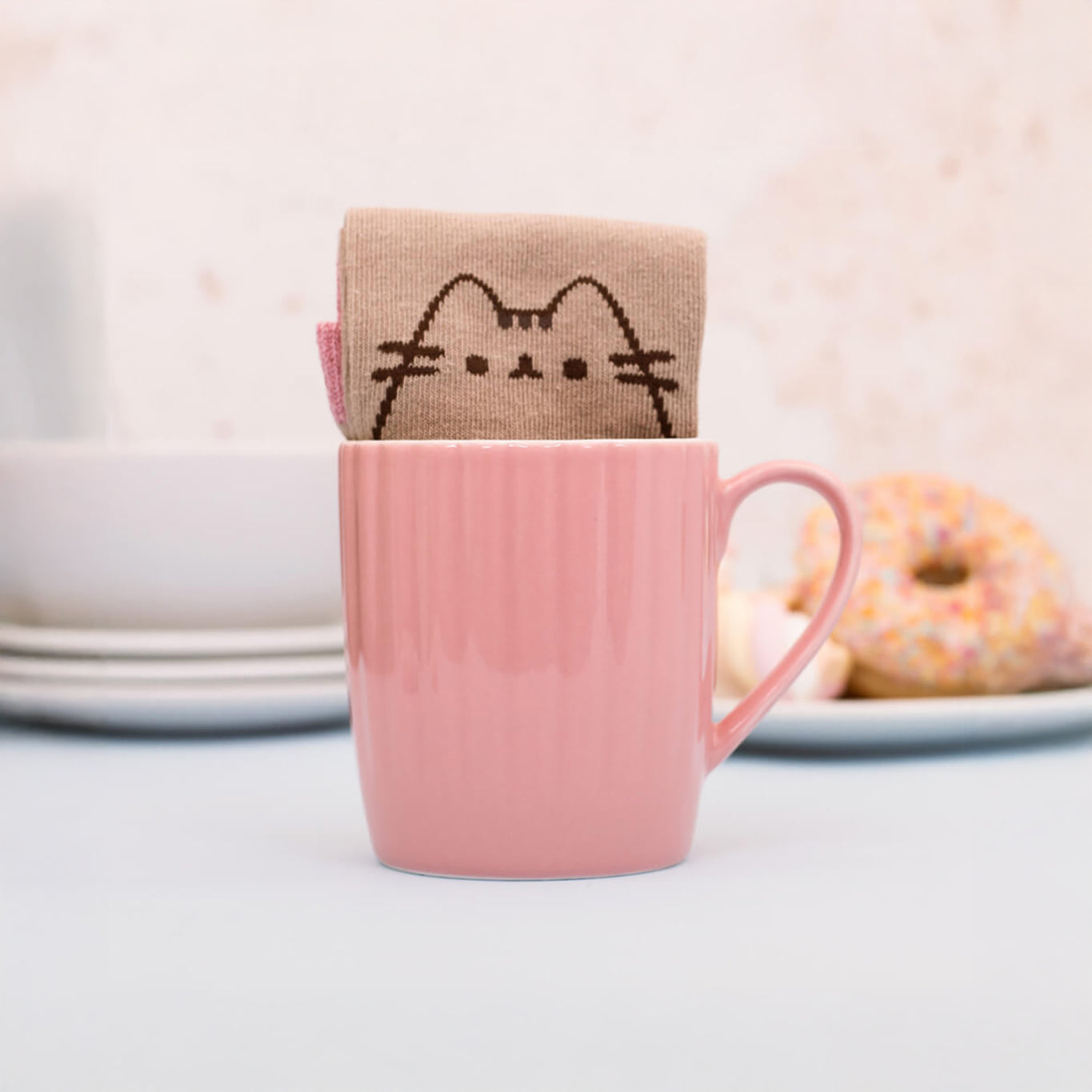 A delightful pink ceramic mug holds a neatly folded beige sock featuring a whimsical cat face pattern, with plates and a donut adding to the cozy atmosphere in the background. This is part of the charming Pusheen Sock in a Mug Gift Set from Pusheen.