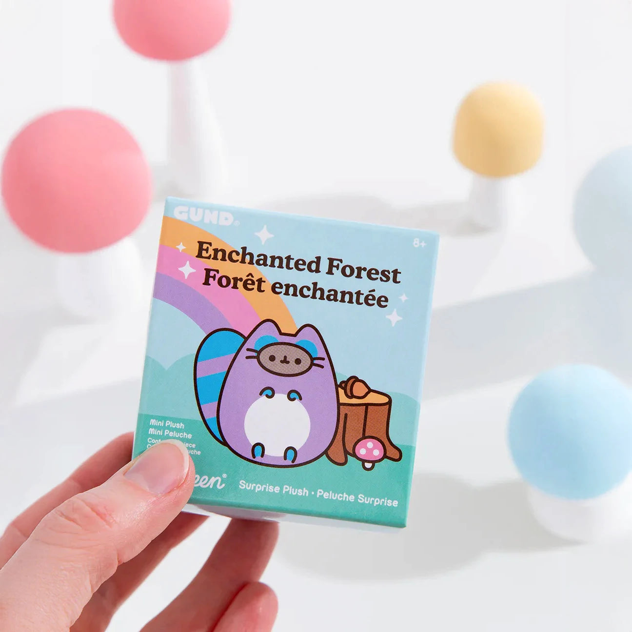 A hand holds a Pusheen Blind Box - Enchanted Forest by Pusheen, featuring a cartoon cat and mushroom design; pastel spheres are softly blurred in the background.