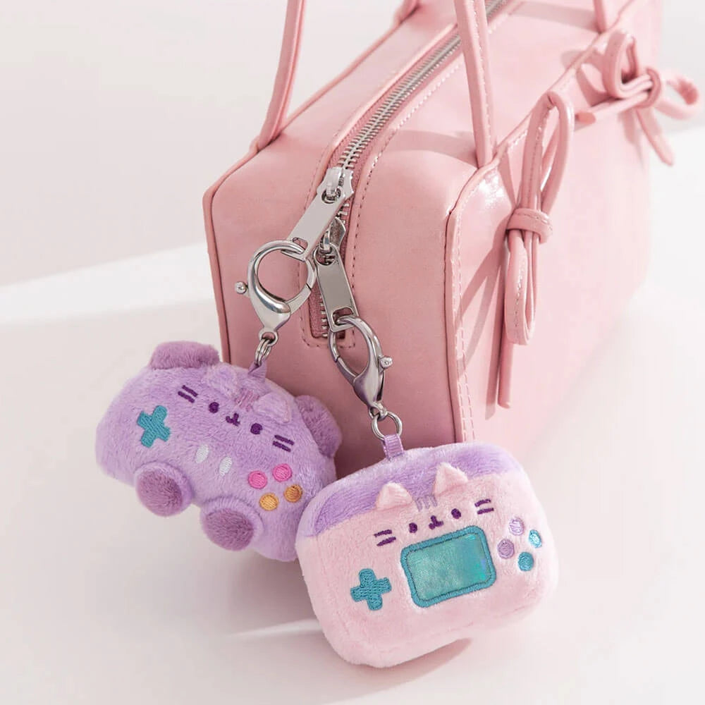 A pink handbag with two pastel Pusheen Blind Box - Gamer Series keychains by Pusheen attached—one shaped like a game controller, the other like a handheld gaming console.