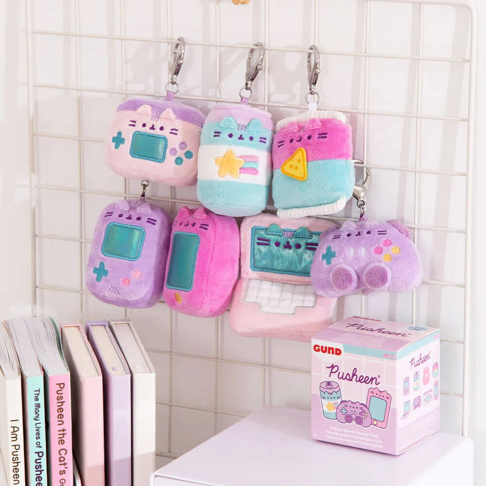 Seven pastel Pusheen Blind Box - Gamer Series plush keychains by Pusheen, each with unique gaming-themed designs, hang on a grid panel above books and a Pusheen toy box—perfect for fans or collectors seeking their next surprise.