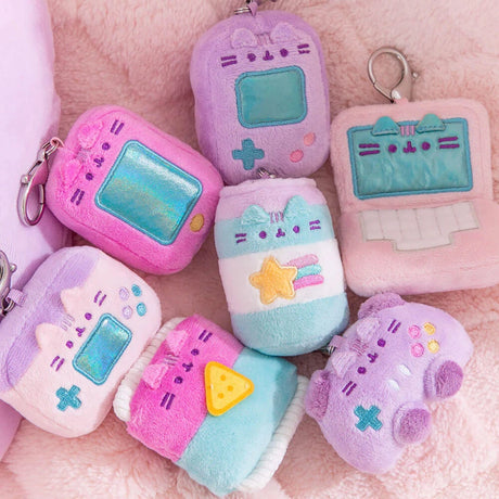 Seven pastel Pusheen Plush Keychains from the Gamer Series, shaped like retro electronics with embroidered cat designs, are arranged on soft pink fabric—each a surprise in the Pusheen Blind Box - Gamer Series collection.