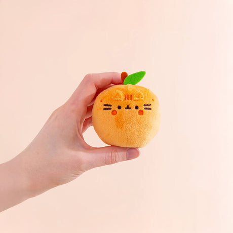 A hand holds a small, round, orange cat plush toy with a green leaf on top, evoking the fruity surprise from the Pusheen Blind Box - Fruits Series by Pusheen.