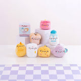 Plush Pusheen cat-themed toys from the Kitchen Surprise Series are displayed on a checkered surface, showcasing designs such as honey, toast, milk, and a pink jar. In the background sits a Pusheen Blind Box from the same series.