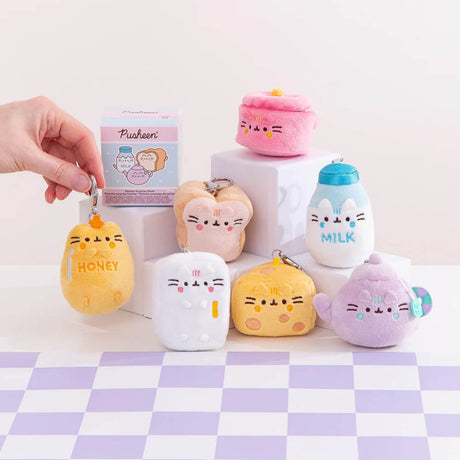 A hand holds a plush keychain featuring a honey theme. Mini plushies, resembling milk, a teapot, and desserts, are displayed on a checkered surface. In the background, there's a box labeled "Pusheen Blind Box - Kitchen Surprise Series" from the Pusheen brand.