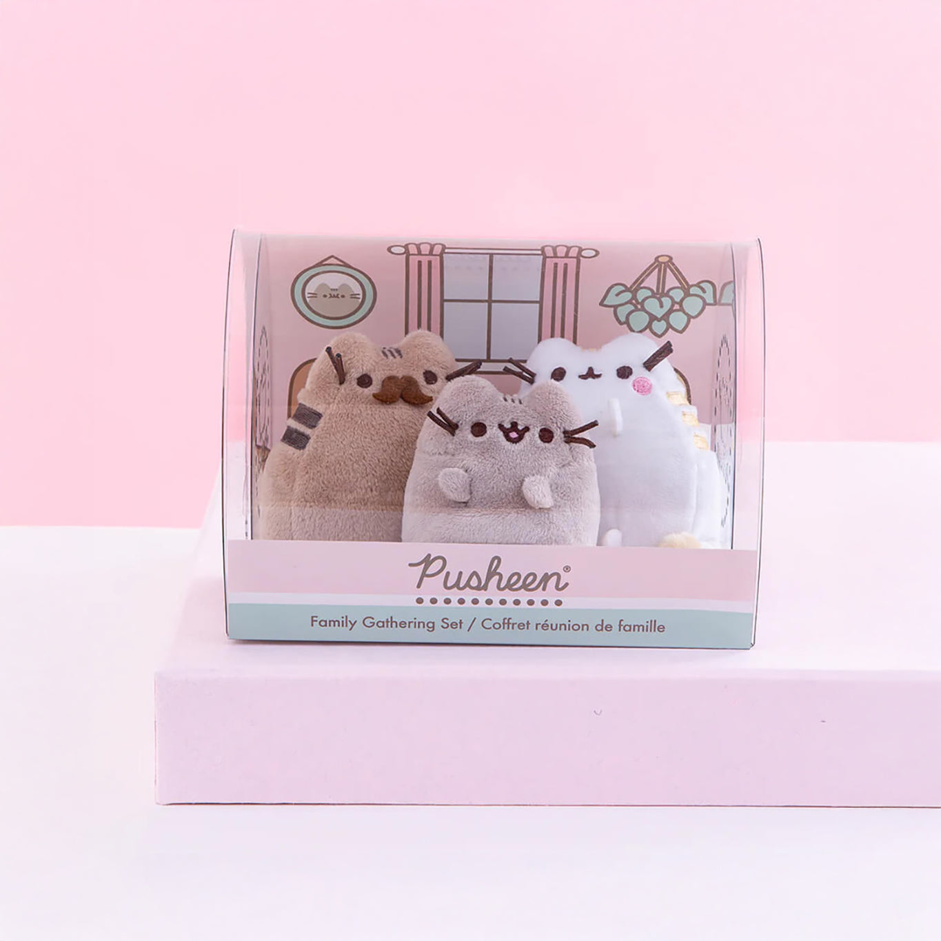 The Pusheen Plush - Family Gathering Gift Set, from the Pusheen brand, includes three plush cat toys beautifully displayed on a pink and white surface with a decorative background inside the packaging. A delightful choice for any Pusheen enthusiast!