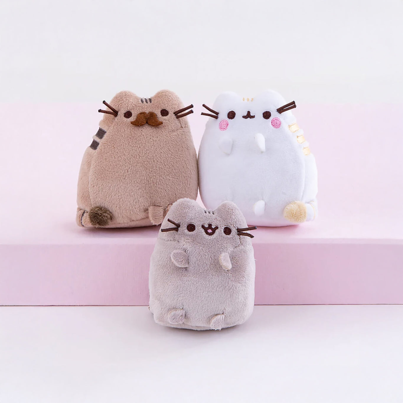 The Pusheen Family - Plushie Gift Set features three plush cat toys on a pink and white platform, ideal for family gatherings. The brown, white, and gray cats each have small whiskers and paws.