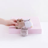 A hand arranges three small plush cat toys from the Pusheen Plush - Family Gathering Gift Set on a pink box against a white background—perfect for fans of the Pusheen brand.