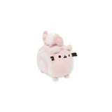 A plush toy from the Pusheen brand, called the Pusheen Plush Mini - Roll Cake, features a pink cube shape with cat details and a swirl pattern on its side. Adding to its charm, there is a small white mini plush rabbit on top.