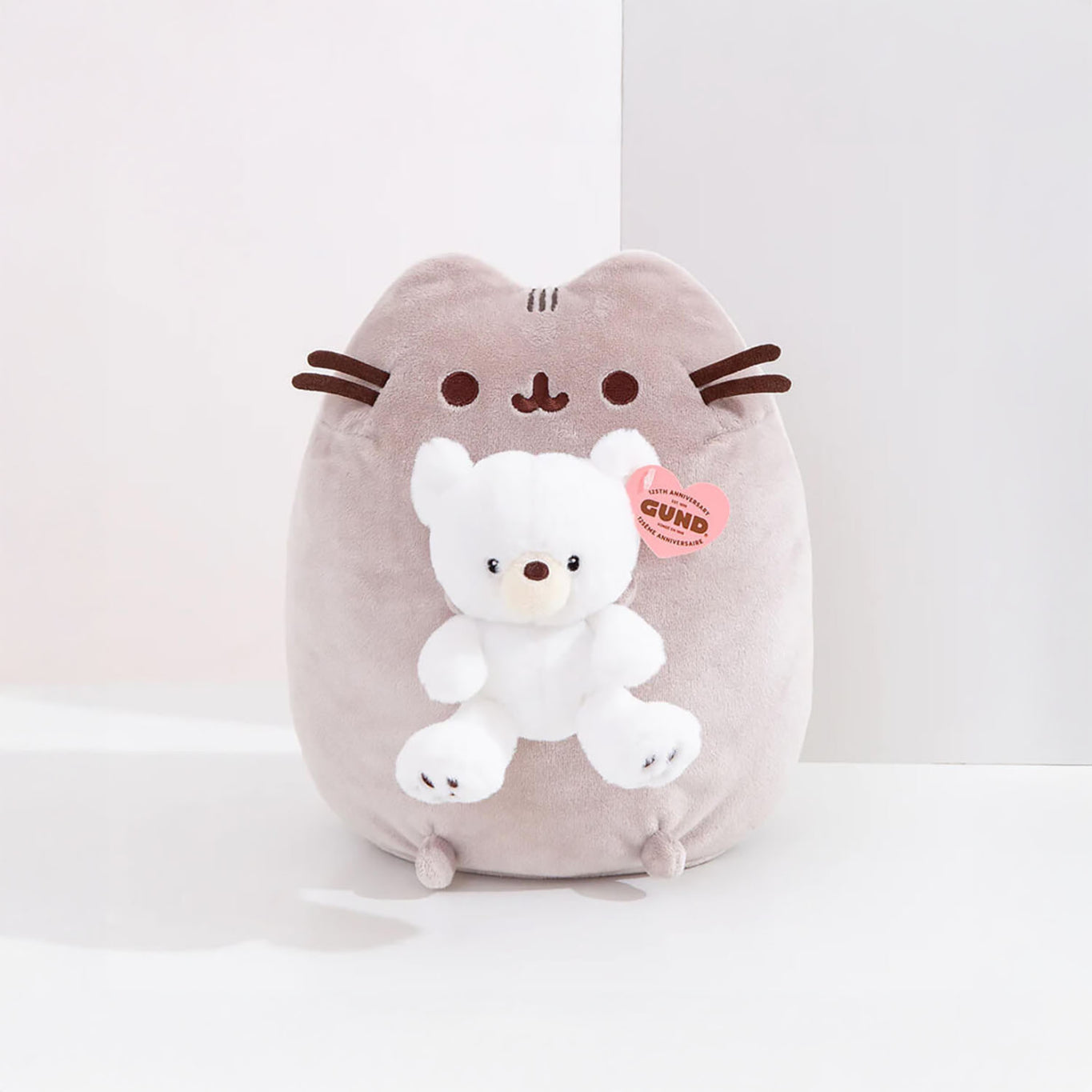 This adorable plush toy, inspired by Pusheen, is shaped like a gray cat and holds a small white GUND bear. It features a unique pink "GUND" tag and is named the Pusheen x GUND Kai Bear Plush - 24cm - Limited Edition.