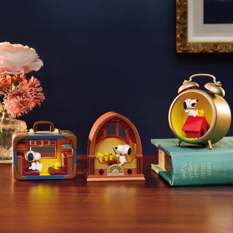 Displayed on a wooden table are three Snoopy-themed figurines, including the Re-Ment Snoopy Blind Box - Antique Diorama. Peanuts fans will appreciate the charm they bring alongside books and a flower vase in the background.