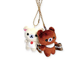 A collectible set featuring Rilakkuma's Korilakkuma mini plush keychain in cream with a red button and a brown bear with a "Rilakkuma" bow, both on gold chains against a plain white background.