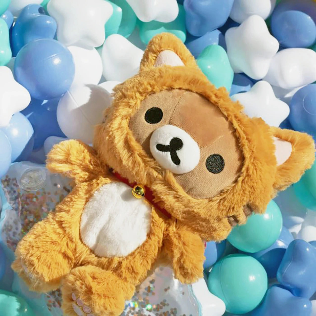 The Rilakkuma Tiger Plush - 23cm by Rilakkuma, dressed in an orange tiger costume, lies among blue, white, and teal star- and sphere-shaped balls—an irresistibly cuddly gift with charming details.
