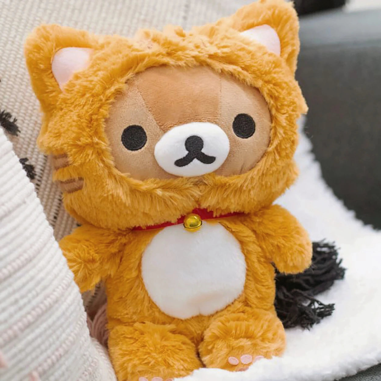The Rilakkuma Tiger Plush - 23cm by Rilakkuma features a cute bear in an orange cat costume with a white belly, sitting on a couch beside a blanket—making it the perfect cuddly gift for any fan.
