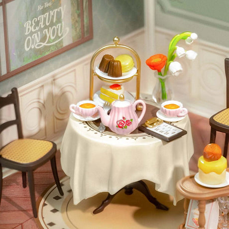 A round table featuring a teapot, two teacups, and a tiered dessert stand captures the essence of the Rolife Sweet Sips Tea Shop - DIY Plastic Mini House. Nearby, two chairs await guests as "Beauty on You" decorates the wall—ready to come alive with intricate details.