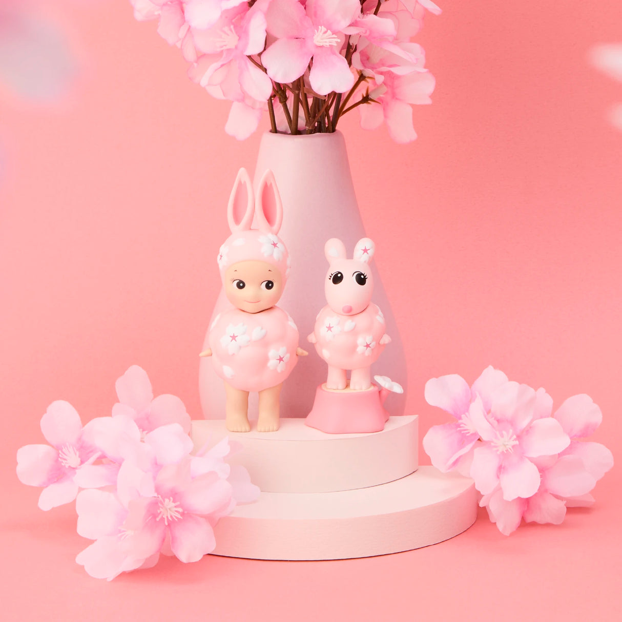 Two Sonny Angel Cherry Blossom Series rabbit dolls stand on white platforms with pink flowers and a vase in the background, all set against a pink backdrop—a charming treasure for collectors.