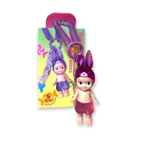 This adorable keychain features a doll with a purple bunny hat, pink skirt, and purple spiral cord. Part of the Sonny Angel Candy Store Series - Blind Box, it includes packaging with a cartoon version marked "5 kinds" for an exciting surprise element.