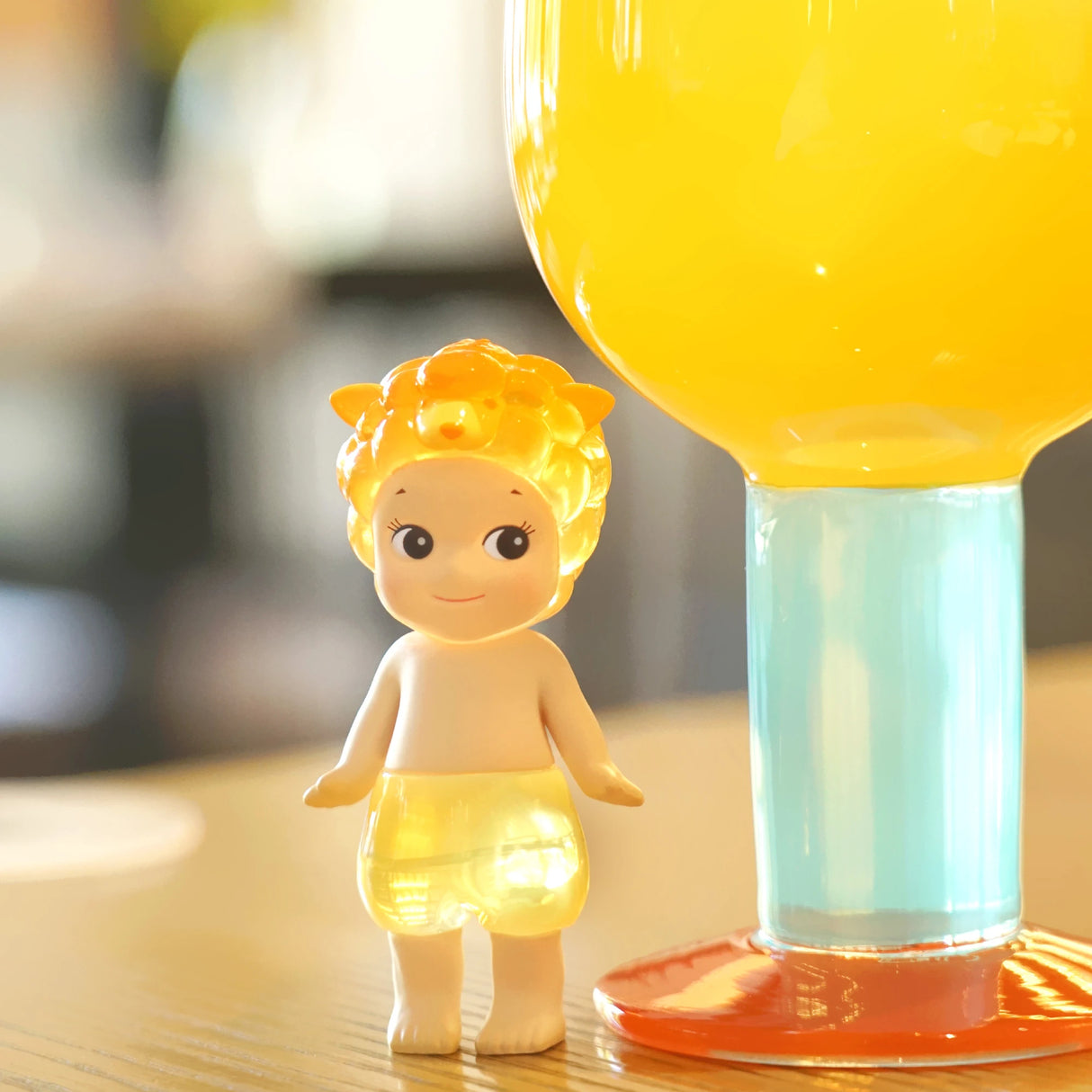 A figure from the Sonny Angel Candy Store Series - Blind Box by Sonny Angel, wearing a lion helmet, is next to a large glass of yellow liquid on a wooden surface.