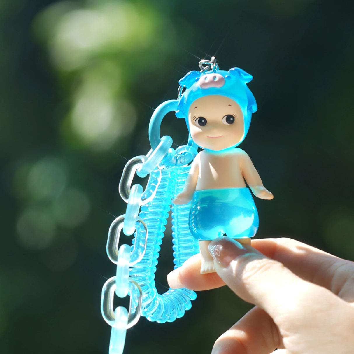 A person holds a Sonny Angel Candy Store Series figure, featuring a blue pig hood and transparent body, attached to a blue spiral keychain. The background is blurred greenery, evoking the surprise of opening a Sonny Angel Blind Box by the Sonny Angel brand.