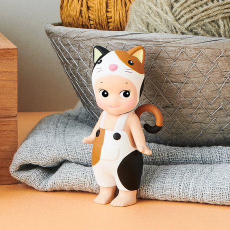 A small figure from the Sonny Angel Cat Life Series by Sonny Angel, dressed in a cat-themed outfit, stands before a basket filled with yarn balls on a textured fabric surface.