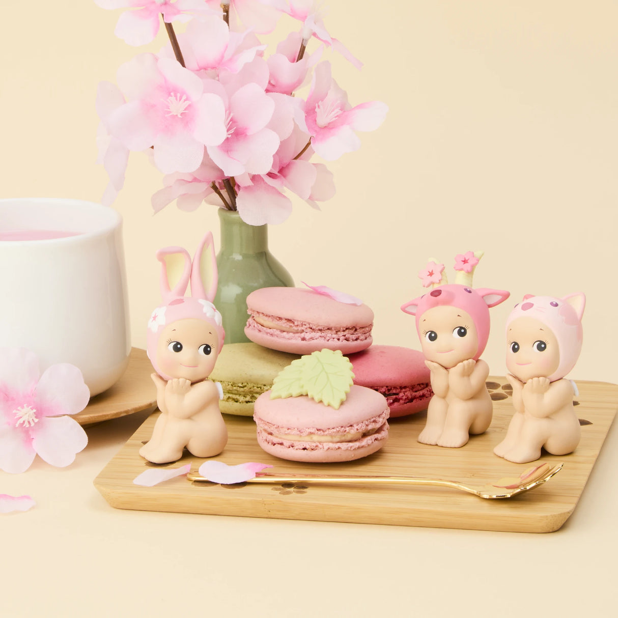 Three Sonny Angel Cherry Blossom Series figurines dressed in animal costumes are placed by pink and green macarons on a wooden board, accompanied by a pink flower arrangement and a cup. This limited edition charm, from the Sonny Angel brand, is perfect for whimsical collectors.