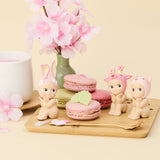 Three Sonny Angel Cherry Blossom Series figurines dressed in animal costumes are placed by pink and green macarons on a wooden board, accompanied by a pink flower arrangement and a cup. This limited edition charm, from the Sonny Angel brand, is perfect for whimsical collectors.