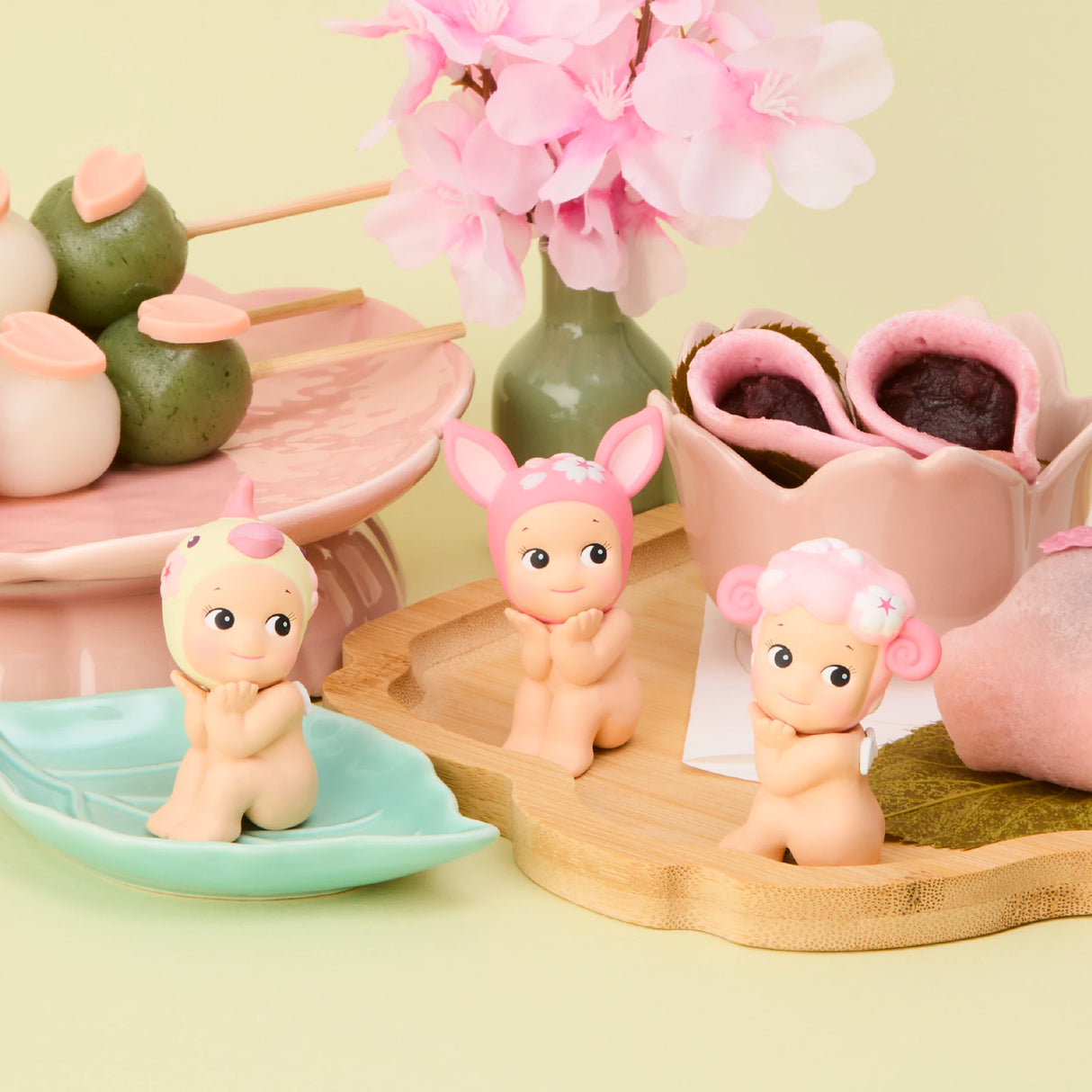 The Sonny Angel Cherry Blossom Series features three charming figurines wearing animal-themed hats, resting among pink and green sweets and sakura blossoms. This limited-edition collection by Sonny Angel is essential for collectors.