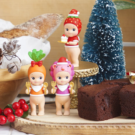 Four festive Sonny Angel Christmas Dinner figurines, each with unique and colorful hats, are arranged on a wooden surface beside a chocolate cake and a small artificial tree, creating a scene reminiscent of a joyful holiday dinner.